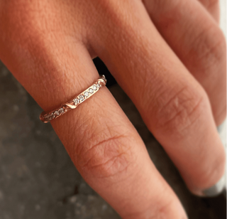 A weathered band in 18k yellow gold is detailed with organic twining vines is studded with colorless brilliant cut diamonds on the top half shown on a finger.