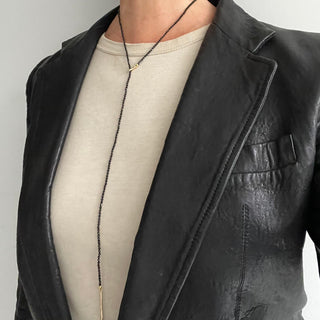 A person in a leather jacket wearing a black spinel gold lariat necklace with a weathered texture closure, featuring a loop and a pull-through bar.