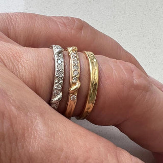 A weathered band in 18k yellow gold is detailed with organic twining vines is studded with colorless brilliant cut diamonds on the top half shown on a finger stacked with a platinum eternity and a gold Everlasting. 
