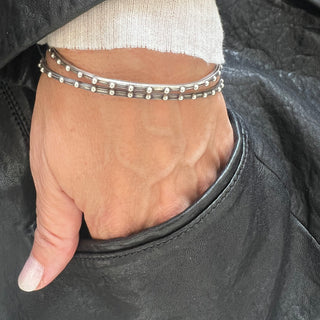 Two silver bangles by Karen Karch, one with a round shape and the other with an oval shape, both featuring a weathered texture and embellished with repeating ball details on a wrist.