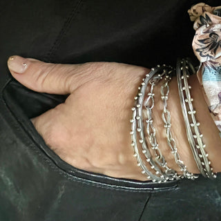 Two silver bangles by Karen Karch, one with a round shape and the other with an oval shape, both featuring a weathered texture and embellished with repeating ball details on a wrist stacked with other bangles and Chain.