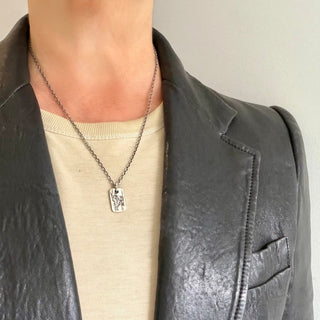 A Karen Karch weathered silver dog tag pendant featuring an embossed fire horse design, shown on a person in a t-shirt and leather jacket.