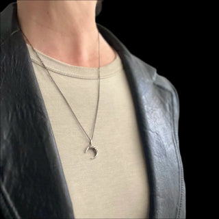A waxing crescent charm necklace with angular metal details, worn by a person with a black jacket and a beige top.