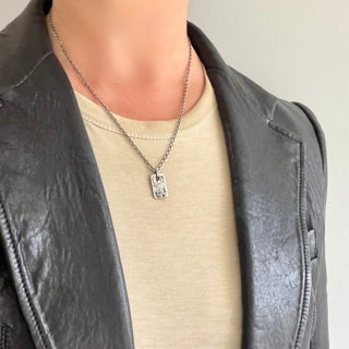 A close-up of a person wearing a silver weathered rectangular dog tag engraved with a dragon on a 20" chain over a beige t-shirt and a black leather jacket.
