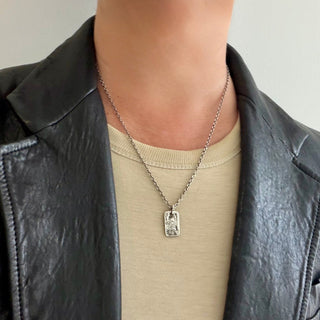 A close-up of a person wearing a silver weathered rectangular dog tag engraved with a dragon on a 20" chain over a beige t-shirt and a black leather jacket.
