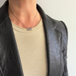 A close-up of a person wearing a silver Karen Karch necklace with the word 'LOVE' spelled out in small letters, styled over a beige t-shirt and a black textured leather jacket.