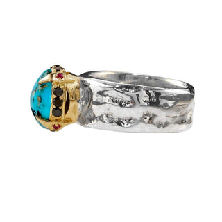 Side view of a Karen Karch weathered silver wide band ring featuring a large round turquoise cabochon raised up in a gold claw setting, studded with Black Diamonds and Rubies around the base shown against a plain white background.
