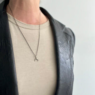 A silver mini waxing crescent charm necklace with angular metal details, worn by a person in a light-colored top and dark jacket.