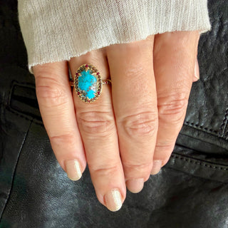 A close-up of a hand wearing a gold Karen Karch Regal Ring with matrix turquoise oval stone with a halo of small cognac diamonds and rubies set against a black leather background and a cream knit sleeve.