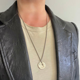 A close-up of a person wearing a silver-Karen Karch necklace with a round weathered tag pendant worn over a beige t-shirt and a dark gray faux-leather jacket.