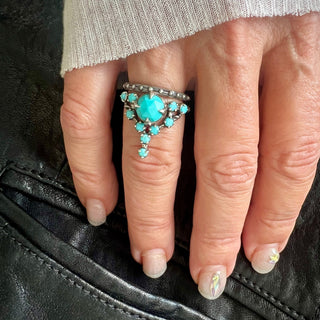 A Karen Karch silver cocktail ring featuring a large round turquoise center stone surrounded by smaller turquoise stones, in a star-like formation stacke with a ball detail silver band on a hand.