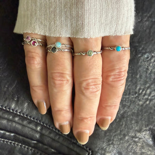A close-up of a hand wearing six Karen Karch silver vine stackers with small colorful gemstones, resting on a black leather surface next to a cream ribbed sleeve.