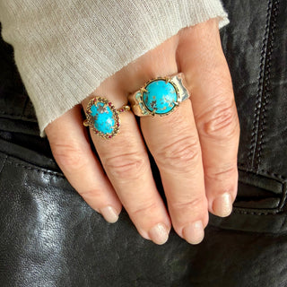 Two Karen Karch silver and gold rings with large turquoise stones worn on a hand, shown against a black leather surface and light beige knit sleeve.