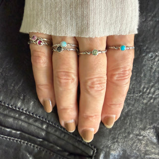 A close-up of a hand wearing six Karen Karch silver vine stacker rings with small colorful gemstones, resting on a black leather surface next to a cream ribbed sleeve.