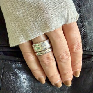 A close-up of a hand wearing two Karen Karch textured silver rings, with a ribbed beige knit sleeve resting above the hand, against a black leather background.
