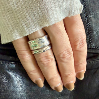 A close-up of a hand wearing two stacked weathered silver rings, resting on a black leather surface next to a ribbed beige sleeve.